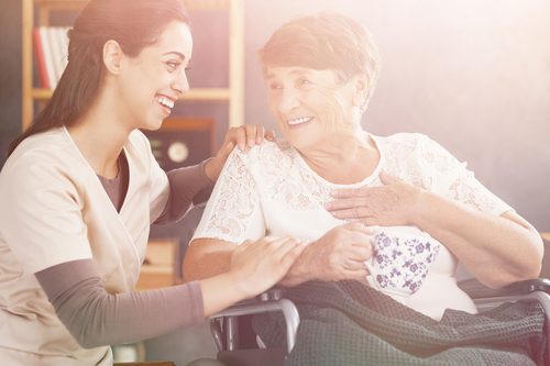 Elderly woman in wheelchair smiling at nurse knelt down behind her